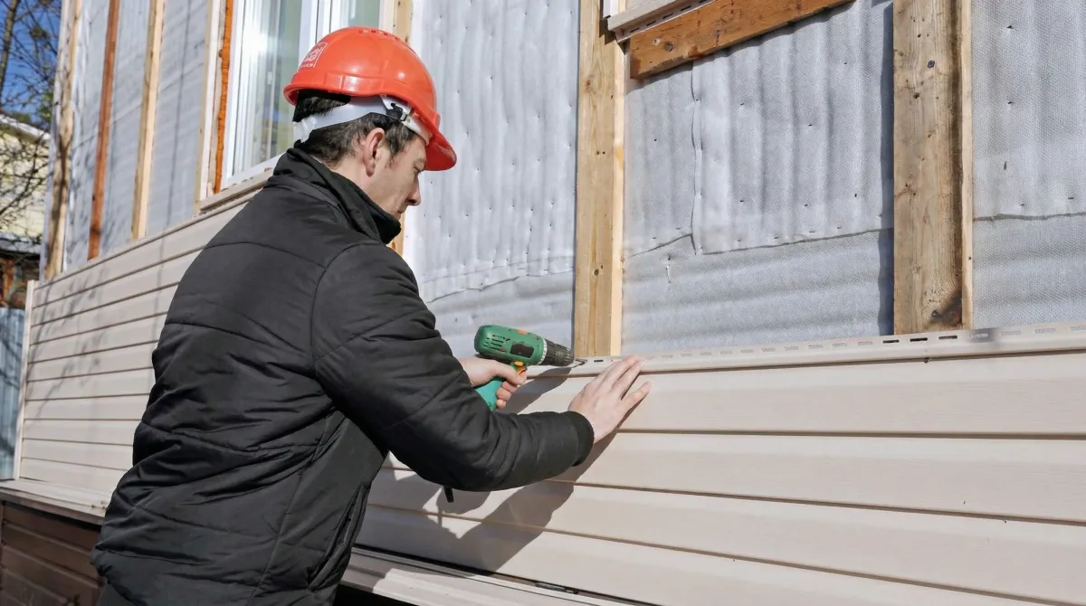 Siding replacement professional using a power drill to install new tan vinyl siding on a home exterior in Omaha, NE